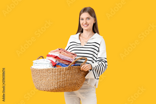 Young woman holding laundry basket with clothes on yellow background