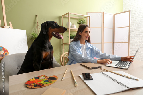 Fotografie Cute Doberman dog near paints palette and beautiful young happy woman working wi