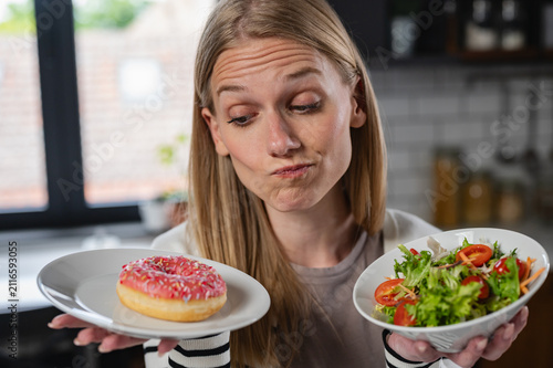 Canvas Print Confused woman deciding between a sugary donut and a healthy salad at home