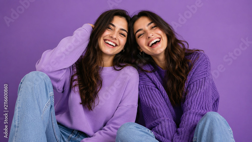 Two smiling women in sweaters pose against a purple background a studio portrait female people friends sister pair laughing joyful happy cheerful positive youthful casual