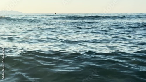 Low angle seascape showing ocean waves advancing toward shore with undulating swells, scattered foam, bright whitecaps, reflective water, and natural coastal movement under daylight