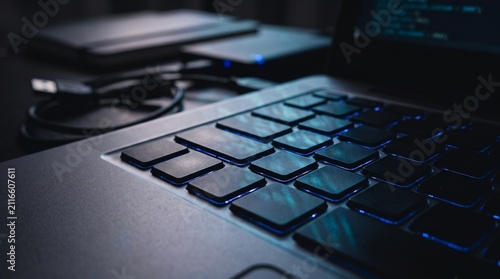 Close up of laptop computer keyboard with code glowing on screen and tech accessories in office