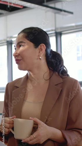 Vertical video: Smiling woman in blazer with beige mug turning head in office as sun causing blink