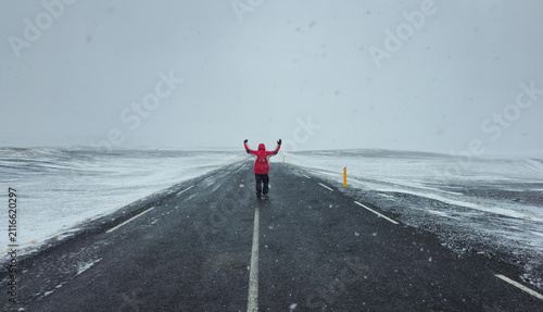 Lone Person in red jacket walking on a long, empty road during a snowstorm, holding arms wide, feeling freedom and adventure. Iceland stormy weather