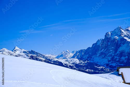A stunning wide-angle winter landscape capturing a pristine, snow-covered mountain slope under a deep blue sky in the Jungfrau region. The photograph highlights a groomed snow trail curving gently acr