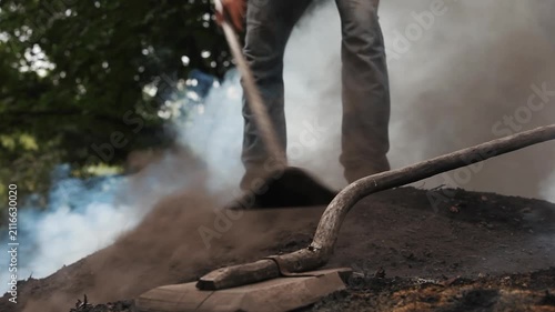 Low angle view of a worker tending a smoking charcoal mound with a traditional tool. Artisanal charcoal production, rural manual labor and traditional craft concept.