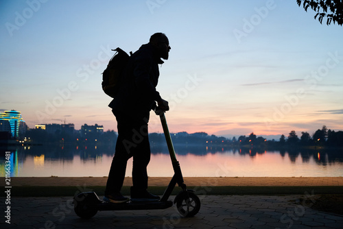 Male commuter on e-scooter near lake at sunset, captured in dark silhouette against glowing skyline, reflections, and tranquil waterfront atmosphere