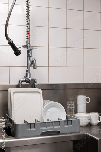 Dirty plates, trays, cups, and drinking glasses loaded into a commercial dishwasher basket in an industrial kitchen.