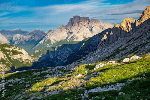 Expansive mountain landscape featuring steep rocky slopes, green summer grass, and distant peaks with sunlight illuminating their rugged forms under a blue sky in the Italian Dolomites