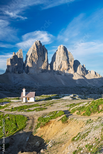 Chapel standing on a hillside with the iconic Tre Cime di Lavaredo peaks rising majestically under a clear blue sky in the rugged Dolomite Alps of Italy