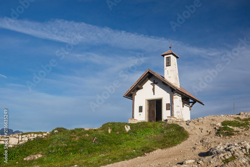 White alpine chapel standing on a grassy hill, a dirt path leading towards its entrance under a clear blue sky, capturing serenity and spirituality. San Briccio chapel Tre Cime di Lavaredo Dolomites