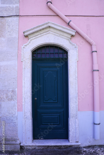 A dark green wooden door with a decorative carved stone frame stands set into a pastel pink building facade in Lisbon, Portugal