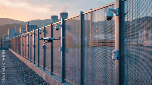 Cinematic overview of integrated secure facility perimeter at dawn. High-tech fence with modern surveillance cameras, sensors, and a blurred security drone in flight. shadows.