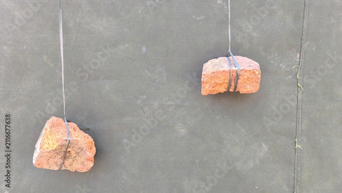 Fotografía Two red brick pieces tied with string used as hanging weights against a dark grey mesh screen