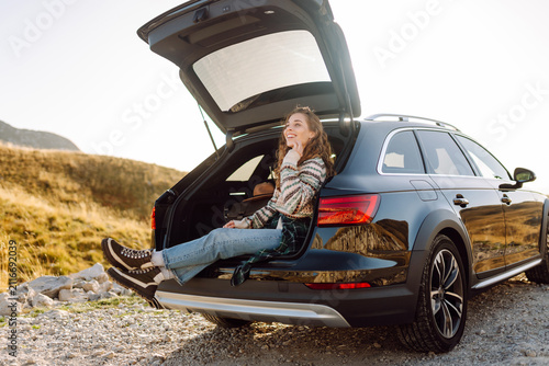 Fotografie Young woman sits in trunk of a car at sunset in the mountains