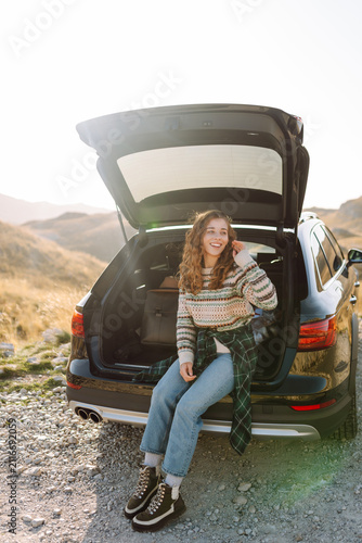 Fotografie Young woman sits in trunk of a car at sunset in the mountains