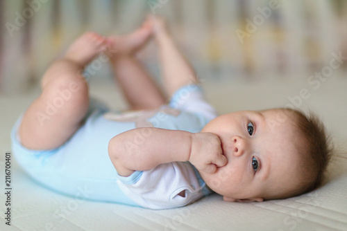 Fotografie A smiling baby lies comfortably on a soft bed in a light blue bodysuit, sucking a tiny fist while looking at the camera
