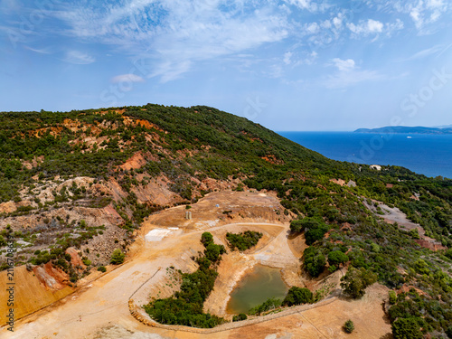 Aerial view of the historic iron mine area near Laghetto delle conche, featuring rugged red geology, sparse vegetation, and a pond overlooking the sea in Rio Marina, Toscana, Italy.