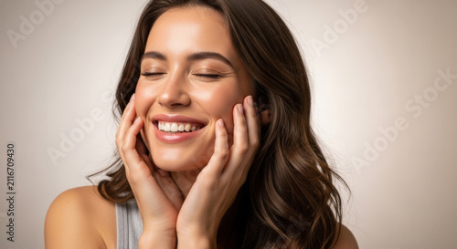 Happy beautiful woman with long brown hair touching her face gently and smiling with closed eyes showing healthy glowing skin against background.