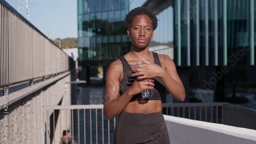 Black African American woman drinks water after an outdoor workout on a modern urban terrace, staying hydrated during fitness training.