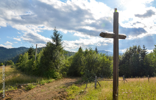 Weathered wooden cross stands in a sunlit meadow beside a dirt path, framed by a green pine forest and a wide blue sky dotted with clouds. Carpathian Mountains, Ukraine