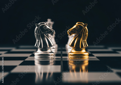 Strategic standoff between gold and silver knight chess pieces on checkered board symbolising corporate competition business challenge plan.