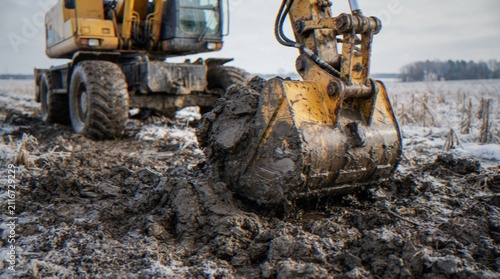 Fotografie A close-up shot of a yellow excavator bucket filled with mud working in a field during winter, with snow patches visible on the ground