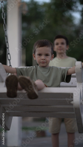Senior brother pushes little brother on outdoor swing in evening light. Happy child smiles on white wooden seat as they share a joyful sibling moment.