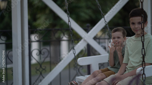 Two brothers swinging on a white porch swing in a quiet outdoor park, smiling and sharing a carefree childhood moment together.