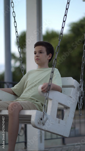 Young boy sits on a white wooden swing in the park at dusk, looking thoughtfully aside with a calm pensive expression amid soft evening light and green trees.