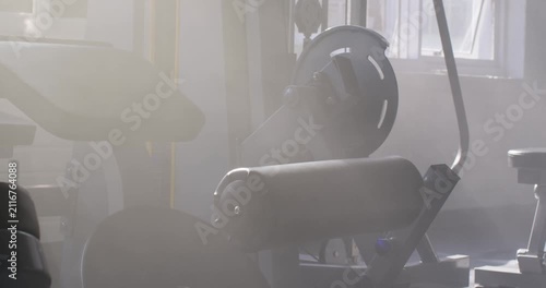 Camera is panning as window light is clearing haze, showing leg curl machine at gym