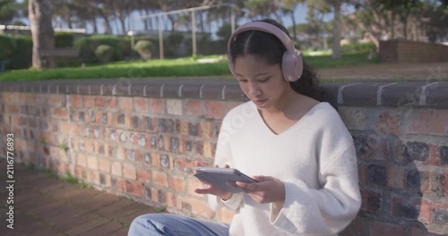 Asian child sitting on brick wall in park with pink headphones tapping tablet, focusing on screen