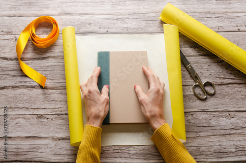 Woman wrapping a book gift with yellow wrapping paper on a wooden table.