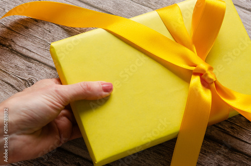 Close-up of a woman's hand holding a yellow gift box with yellow ribbon on a wooden table.