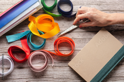 Crafting colorful ribbons, wrapping paper rolls, a gift book and a hand holding scissors on wooden table. 