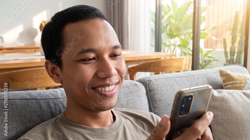Smiling happy young Indonesian man holding cell mobile phone using smartphone sitting at home on couch, scrolling social media, checking financial apps, buying online, texting messages. 