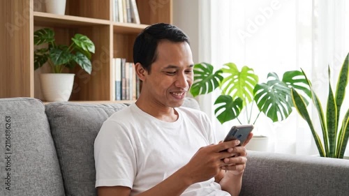 Smiling happy young Indonesian man holding cell mobile phone using smartphone sitting at home on couch, scrolling social media, checking financial apps, buying online, texting messages. 