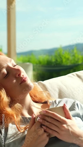 Peaceful redhead woman in pajamas enjoying her morning coffee on a sunny day, relaxing on the couch and looking thoughtfully at the beautiful view from her home, feeling calm and content