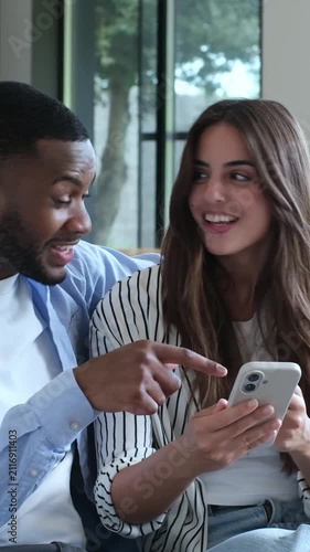 Excited young interracial couple looking at a smartphone screen, celebrating good news, an online win, or a successful achievement together while sitting on the sofa at home