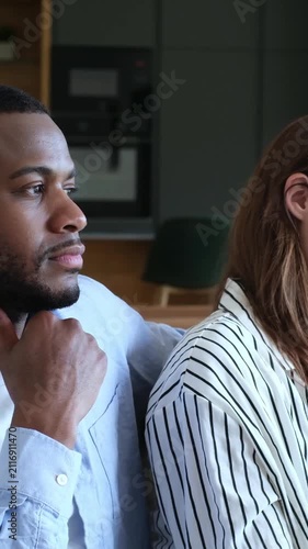 Unhappy and resentful interracial couple sitting back to back on the sofa, ignoring each other after a difficult fight, symbolizing a relationship crisis, misunderstanding, and conflict