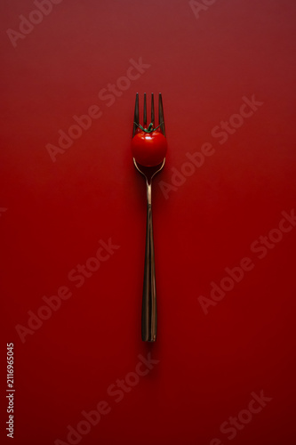 фотографія A single fresh cherry tomato impaled on a metal fork against a minimalist red background