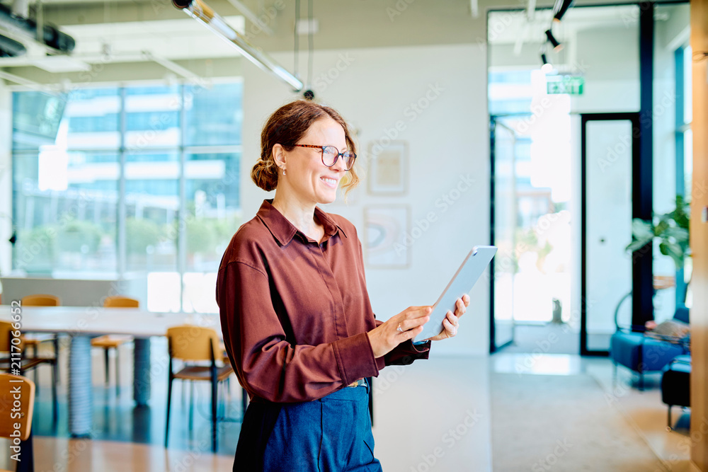 Fototapeta premium Smiling mid adult businesswoman holding a digital tablet. She is standing in a modern office, engaged with technology and work