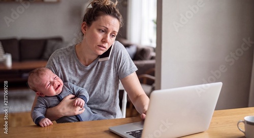 An overwhelmed young mother trying to work on her laptop and talk on the phone while holding a crying baby in her living room.