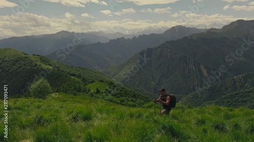 Male hiker climbs an inclined alpine hill using trekking poles, with stunning layers of mountain ridges, green meadows, and soft summer light creating an inspiring outdoor scene.