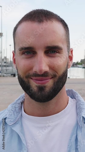 Vertical HD video portrait of smiling young Spanish man looking at camera in Barcelona. Confident millennial standing outdoors on a sunny day. Lifestyle and confidence concept.