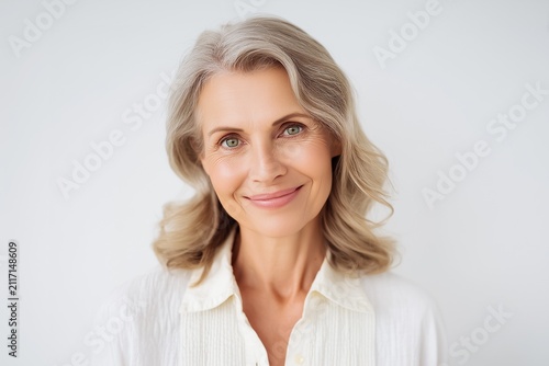 Studio headshot portrait of smiling mature senior woman with long wavy grey hair against plain light white background
