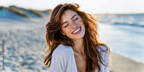 Happy young woman laughing with closed eyes and long wavy windblown hair enjoying, standing, walking on sand beach coast near ocean, sea waves with summer vacation sunlight, in nature