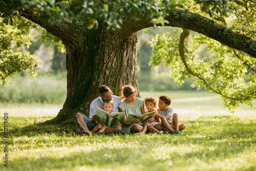 family reading storybooks together under a large oak tree in a peaceful park, quiet bonding moment, green surroundings, relaxing outdoor experience, soft natural lighting