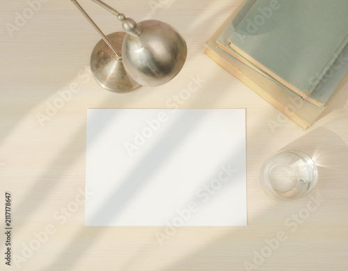 Clean white blank paper sheet lies on a light wooden desk, surrounded by a metallic lamp, old books, and a glass of water, all bathed in warm natural sunlight creating interesting shadows