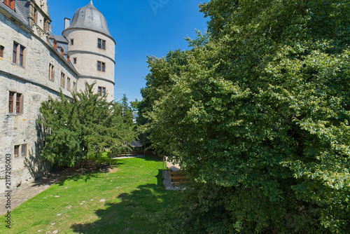 Foto A scenic view of Wevelsburg Castle, showcasing its stone walls and round tower against a bright blue sky, framed by green trees and a grassy lawn